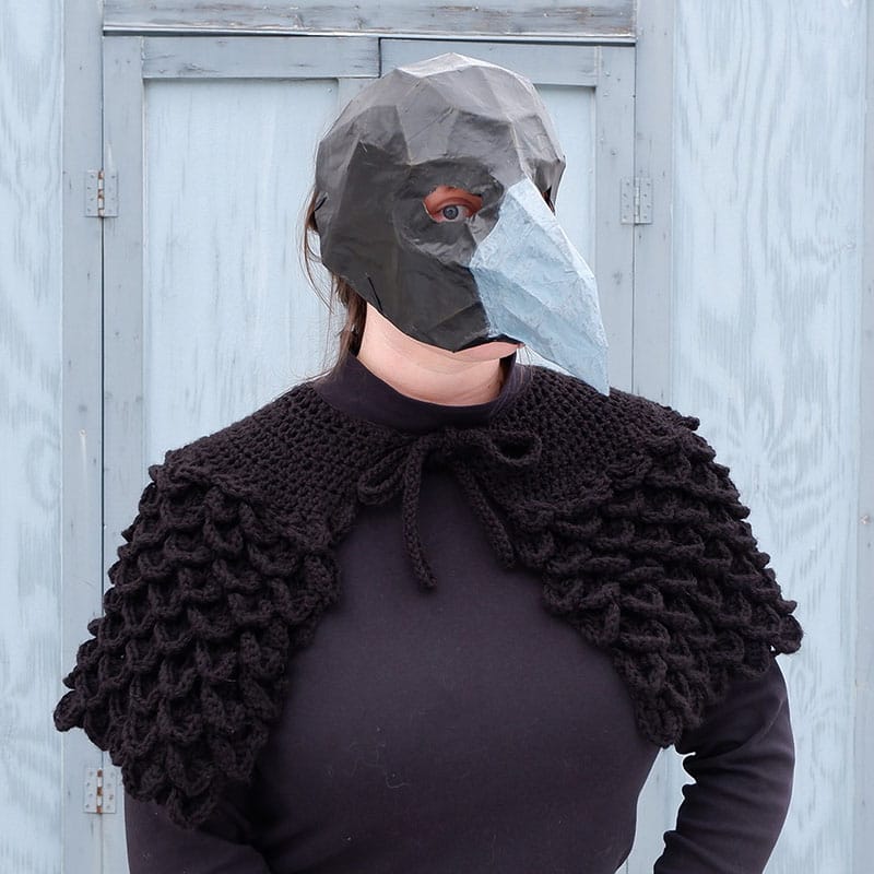 Portrait of a woman in a papier-mâché bird mask wearing a crochet shawl that gives the effect of feathers.