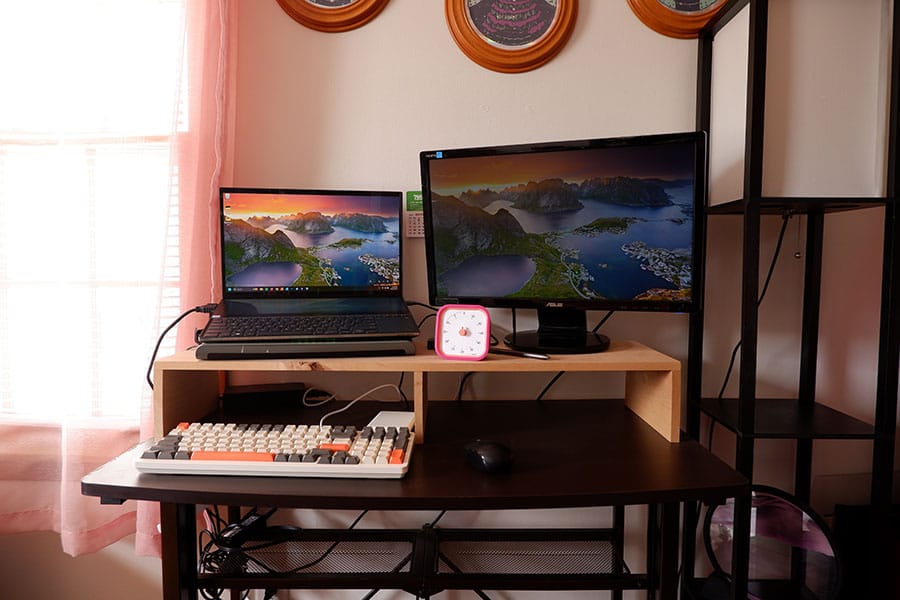 Photo of two laptops elevated on a wooden platform. There's a mechanical keyboard in front and a mouse. Above are three circular prints that are cut off at the top. There's a window to the left and a lamp to the right.